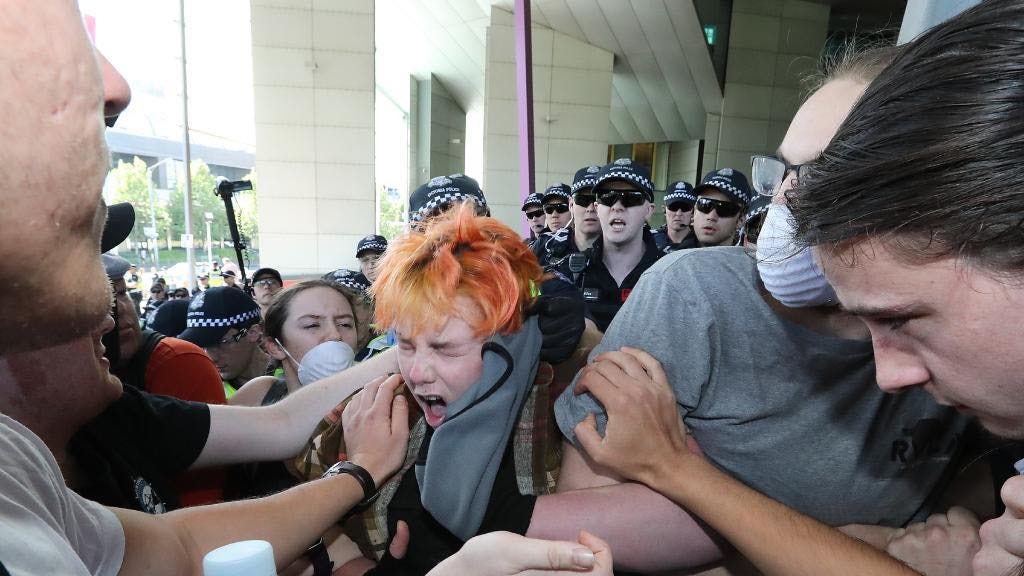 A young orange-haired person in a crowd, cops close behind but being pushed back, with face screwed up and hair mussed, both freshly bared by the half-hidden cop with a gloved fist still clenched around a balaclava