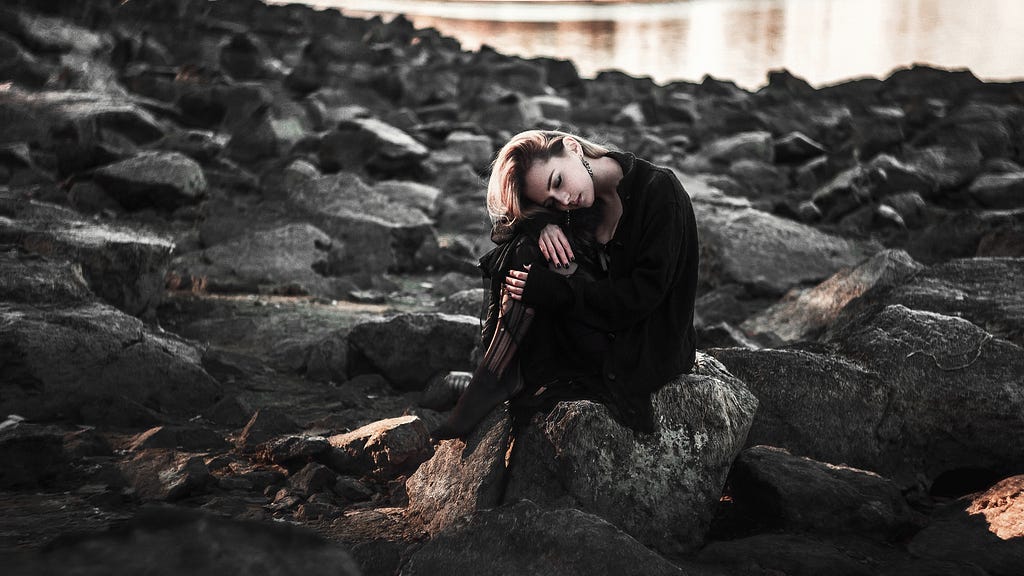 Broken and Sad girl sitting beside a lake on some rocky boulders.