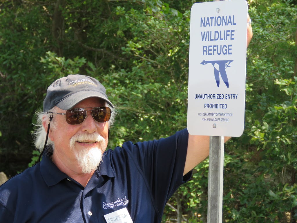 Man in baseball hat and glasses standing next to a sign that reads National Wildlife Refuge Boundary