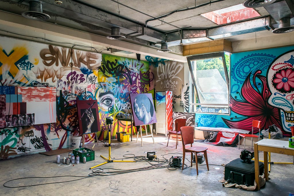 A corner view of an art studio with grafitti-style work on the walls, chairs and equipment strewn about, and natural light flowing in from a slightly open window and sky light.