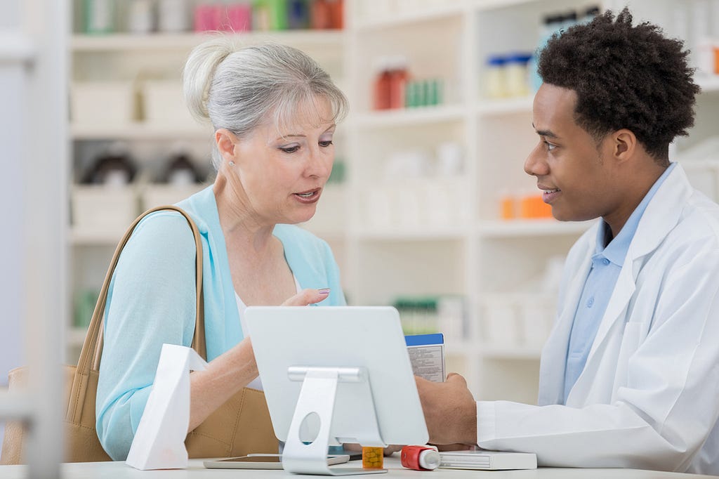 A senior woman stands at the pharmacy counter with a male pharmacy technician and asks about her medication