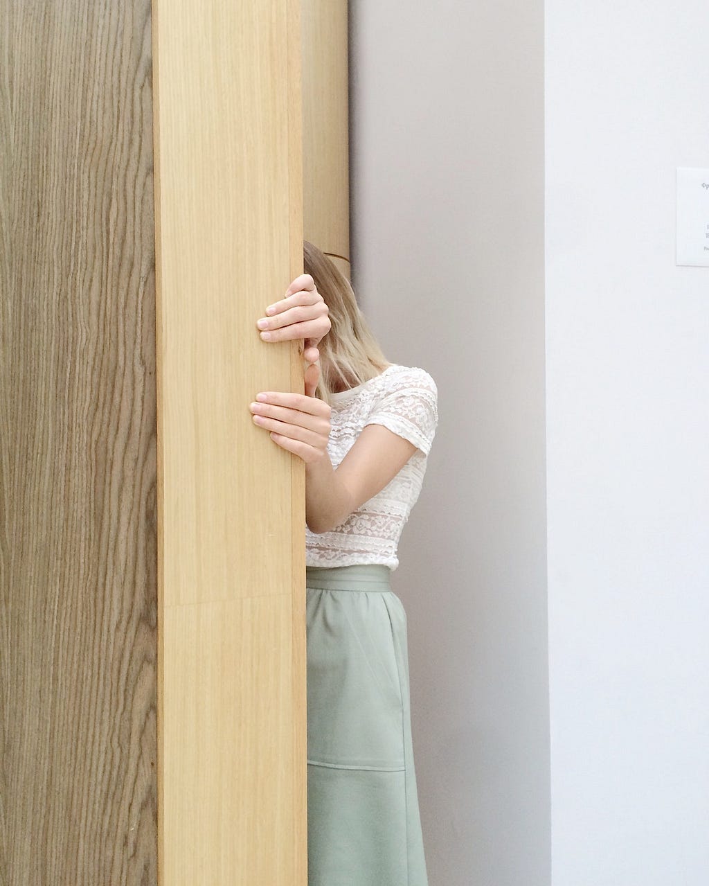 Woman in a white lace short-sleeved top and a light green skirt hiding behind a brown wall