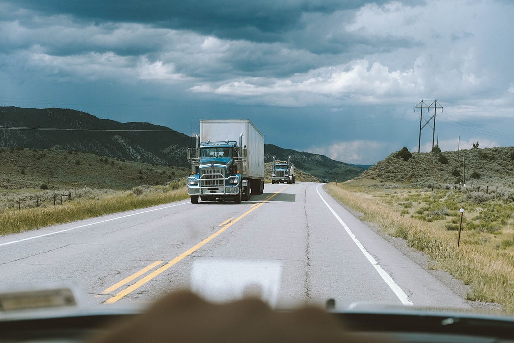 Two semi trucks drive down a highway to deliver their shipments while a car watches them.