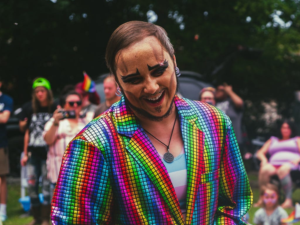 A drag queen smiling while wearing a rainbow-checkered sports jacket & a blue, white, & pink striped shirt — possibly representing the transgender flag — underneath. There is a crowd of people behind them, watching the show