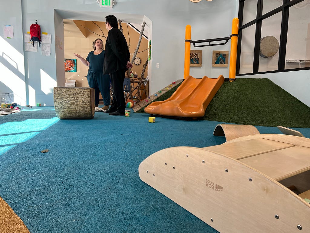 Two people stand and talk inside an interactive play room at a child care facility.