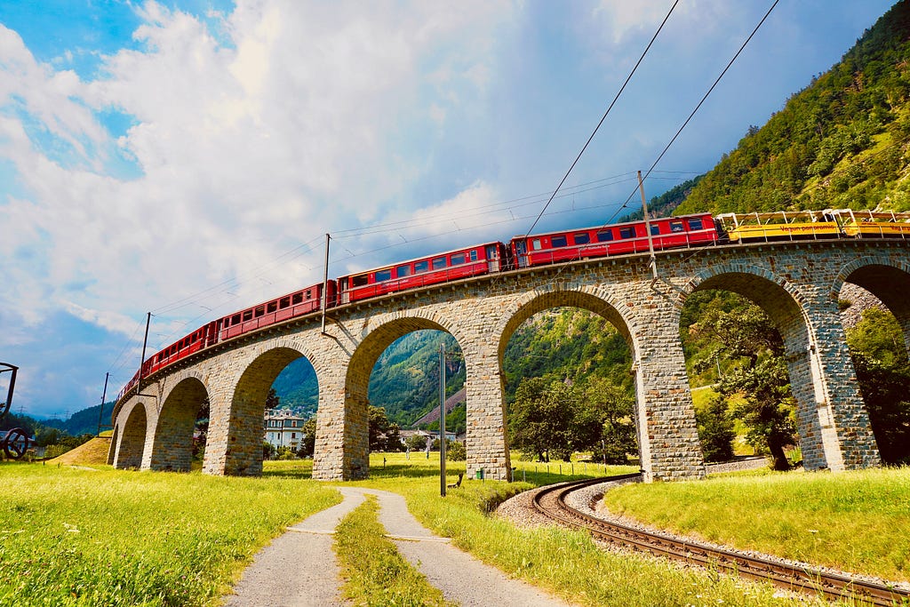 a train on a raised trainway consisting of arches. One train track runs beneath one arch, and a dirt road underneath another. There is a mountain behind the raised trainway and the sky is visible with some clouds.