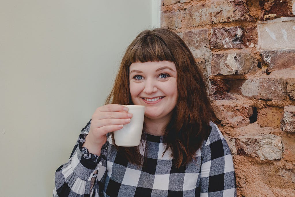 Author Lauren Aitchison standing in front of an exposed brick and white wall, wearing a checkered jumper, and holding a cup.