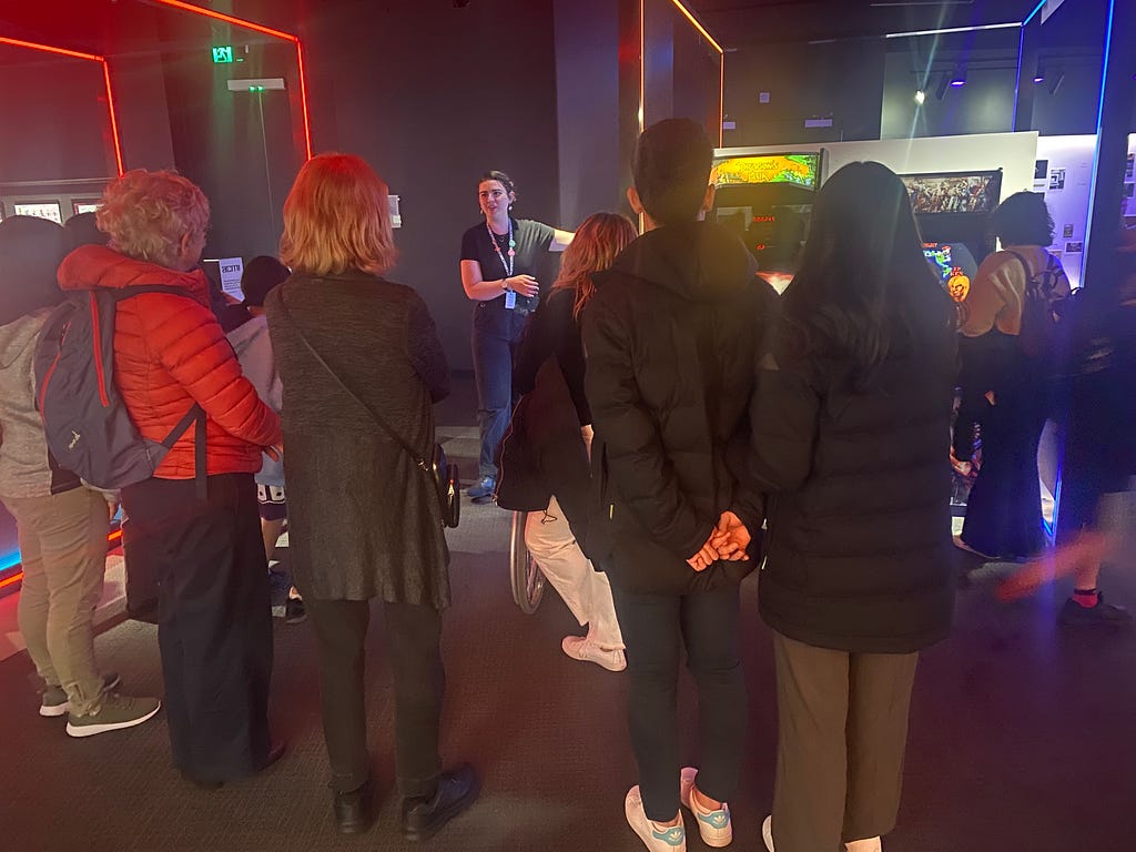 An ACMI VX guide talking to a group of people gathered around an arcade game inside the ACMI exhibition ‘Story of the Moving Image’