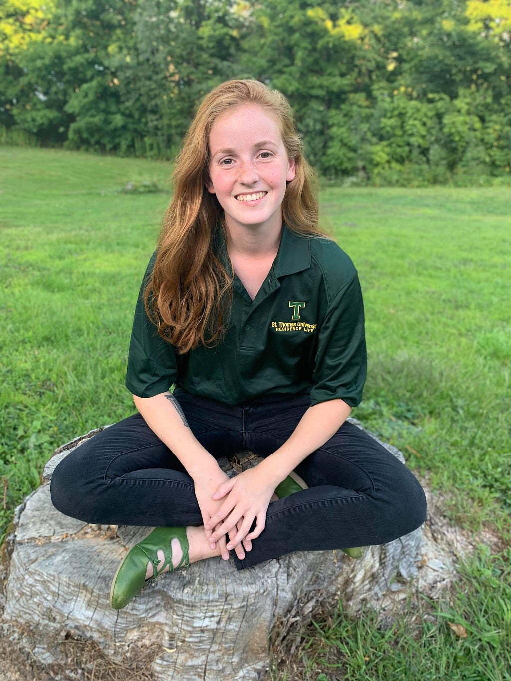 Fiona sits crosslegged on a tree stump outdoors in the summer smiling at the camera.