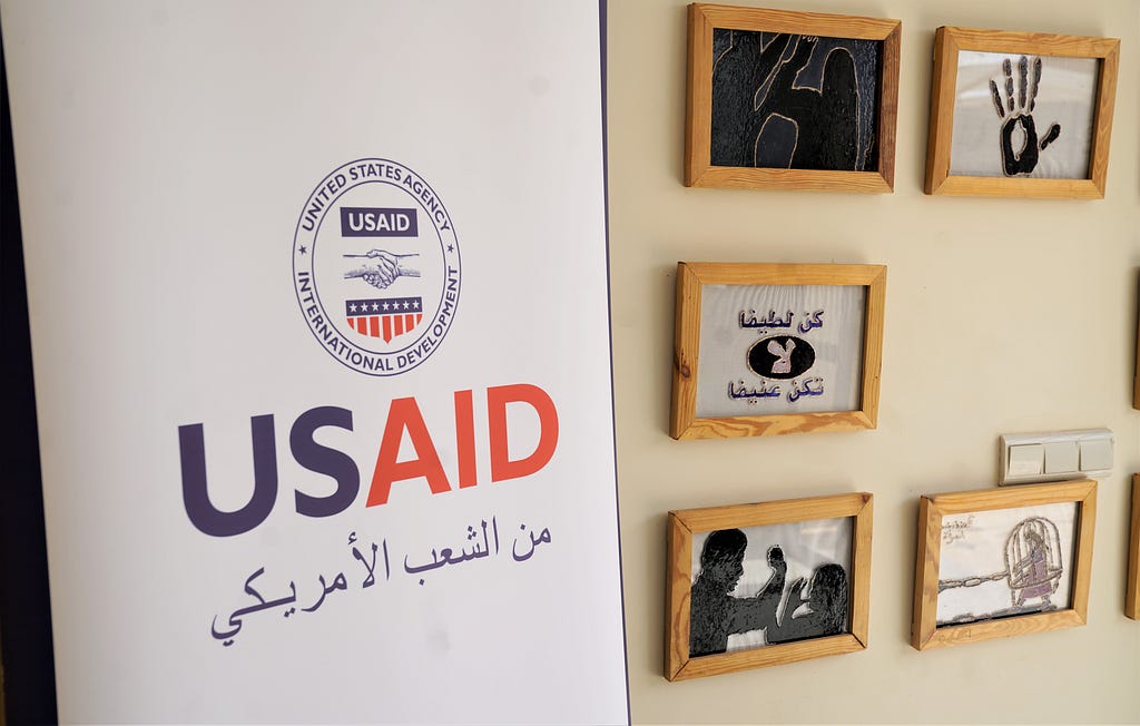 The USAID logo next to five framed art pieces depicting artistic representations of violence against women, including a woman in a bird cage and a hand in the “stop” gesture.