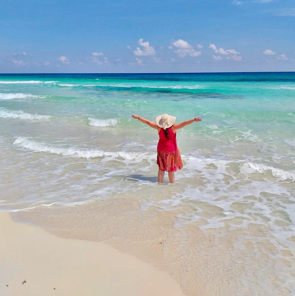 A woman in red wearing a huge beach hat standing ankle-deep in crystal clear Caribbean water with arms stretched wide in complete freedom.