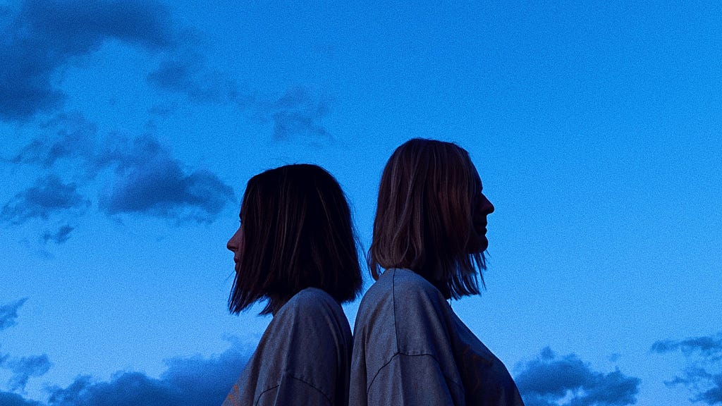 Two white girls with shoulder-length hair in grey shirts sitting back to back in front of a blue sunset background
