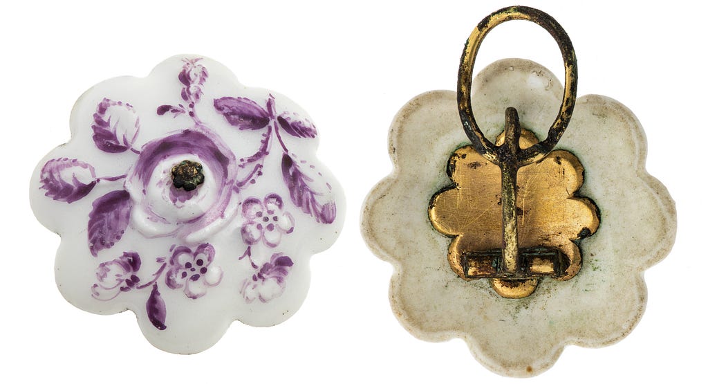 Two earrings, one seen from the front and one from the back. The front is a painted purple floral pattern on white porcelain. The back is a metal fastening clip.