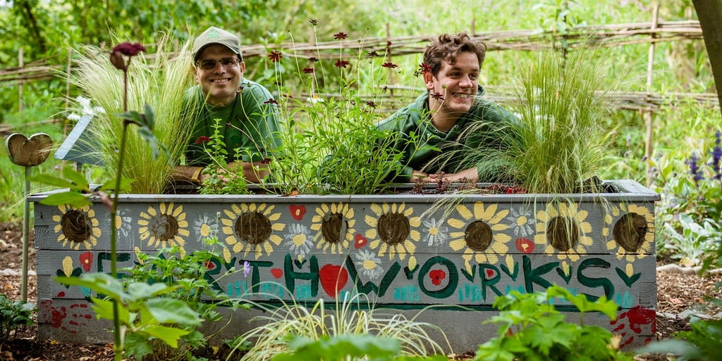 Two white men from Earthworks, wearing green tshirts and leaning over a sign that says ‘Earthworks’ with sunflowers painted on it. One man is wearing a green cap. They are surrounded by lots of green plants.