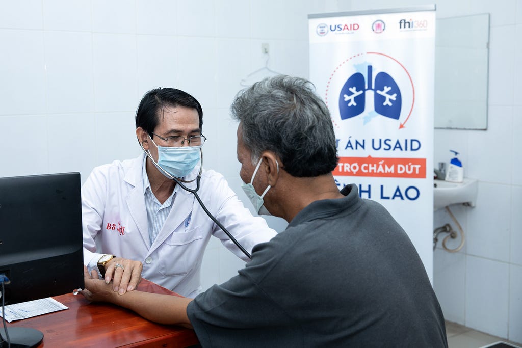 A health care worker listens to a patient’s chest with a stethoscope while also measuring his pulse on his left wrist.