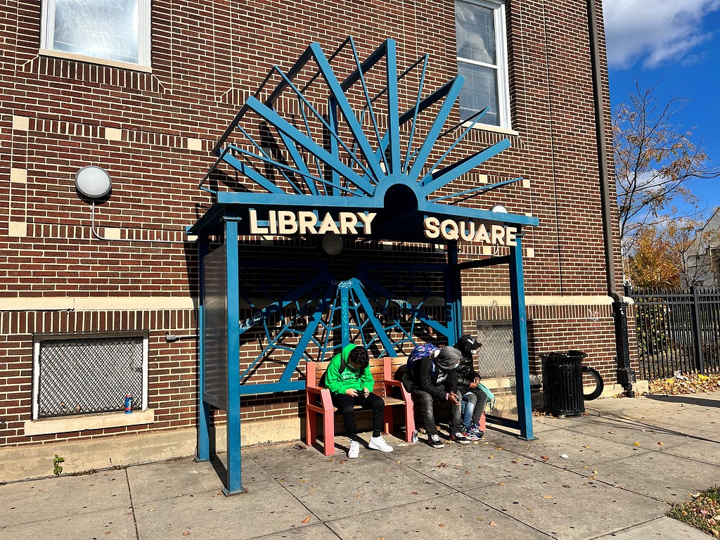 Three young people sit on a bench at a bus stop in Baltimore and look in the direction the bus should come from.