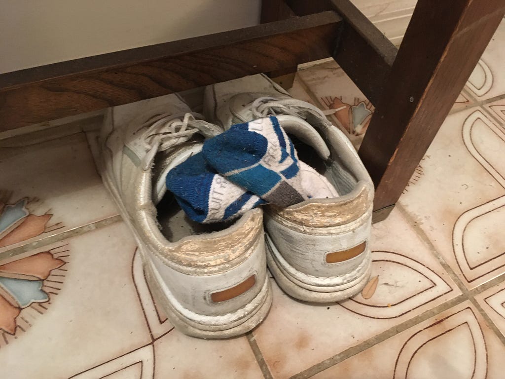 Color photo of men’s white sneakers with socks tucked under a hallway table.