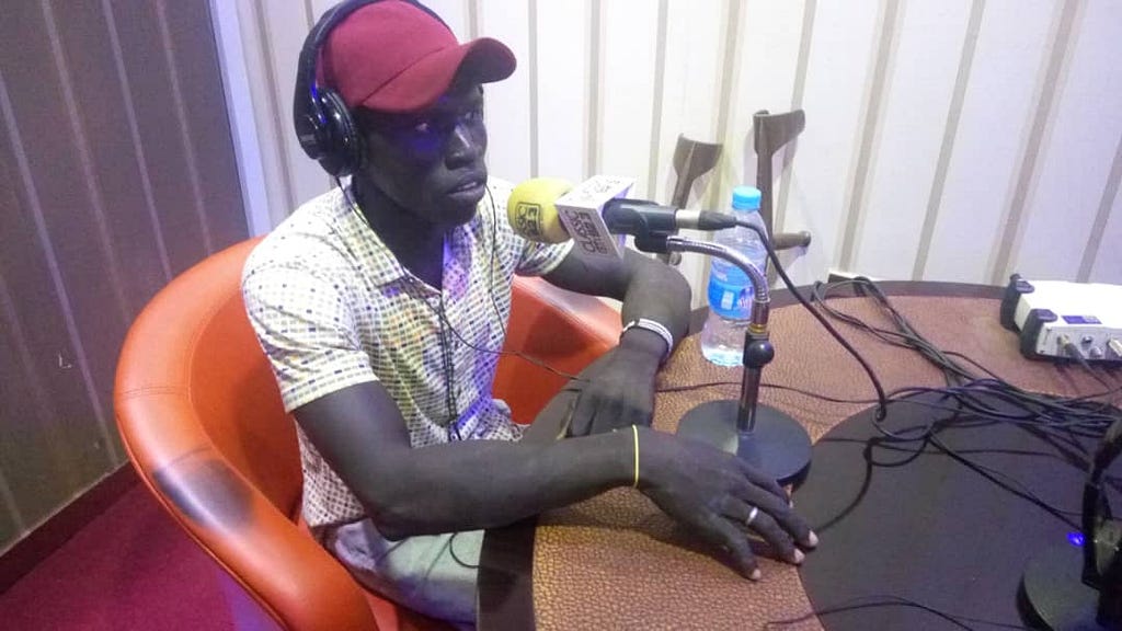 Seme Lado in a radio station talking to a microphone. He is wearing a white checkered shirt and a red hat.