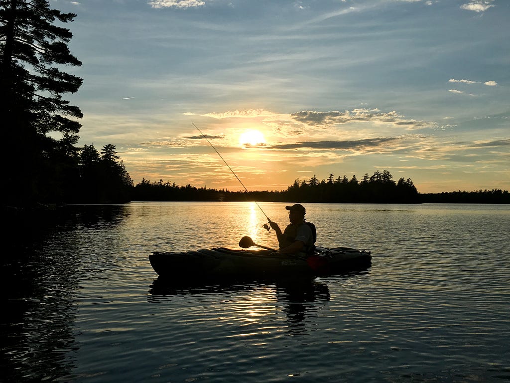 A silhouette of a man in a kayak. The man is holding a fishing rod while floating on a lake as the sun sets in the background.