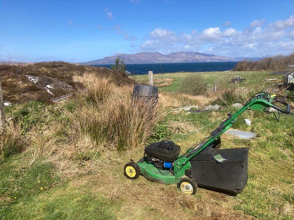 A green lawnmower is sitting idle on wild grassland. There is a stretch of blue open water in the distance, and hills behind