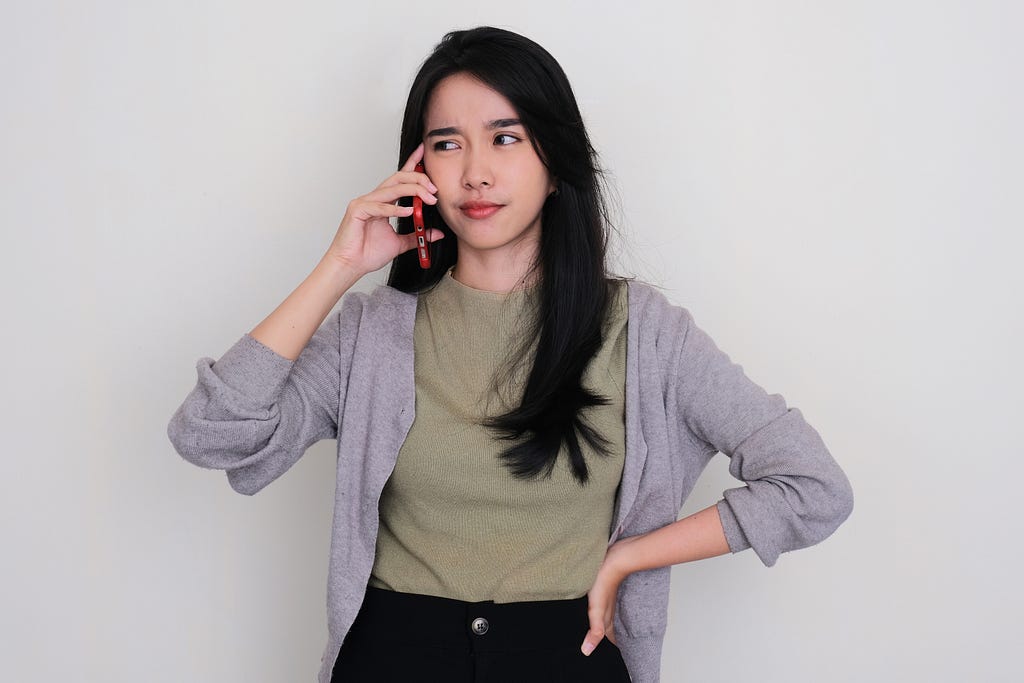 An Asian woman standing with her left arm akimbo holding a mobile phone with her right hand against her right ear. She is listening to someone speaking to her on the phone with a look of disbelief.
