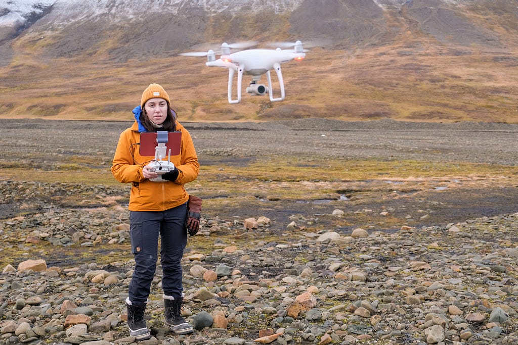 Jess Todd in an orange winter jacket holds a remote controller to operate a white drone, which hovers a short distance in front of her in the air.