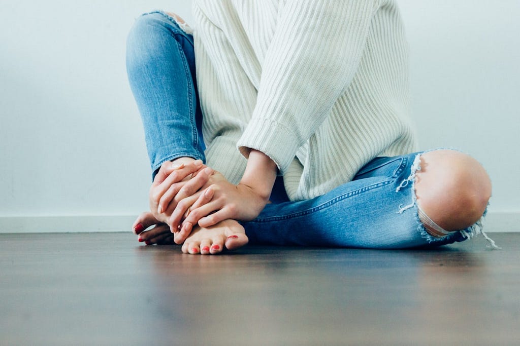 Imani Bahati took this photo, from the chest down, of a woman in a white shirt and jeans with a hole in the left knee, sitting on a hardwood floor.