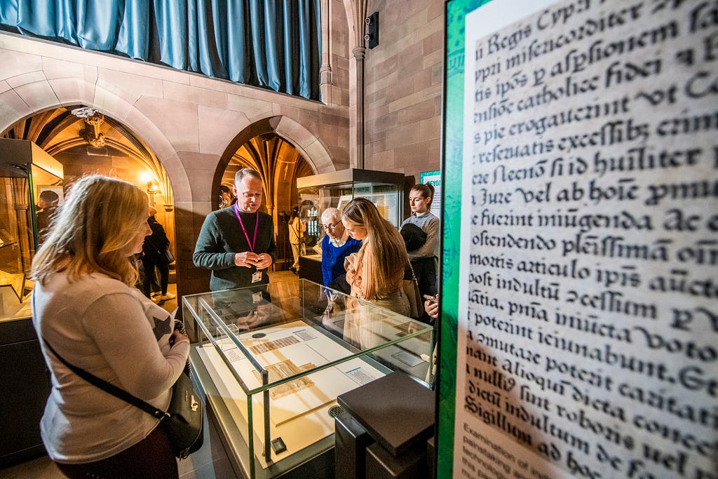 Visitors standing in the Transitions in Print exhibition, listening to a tour and examining the display cases.