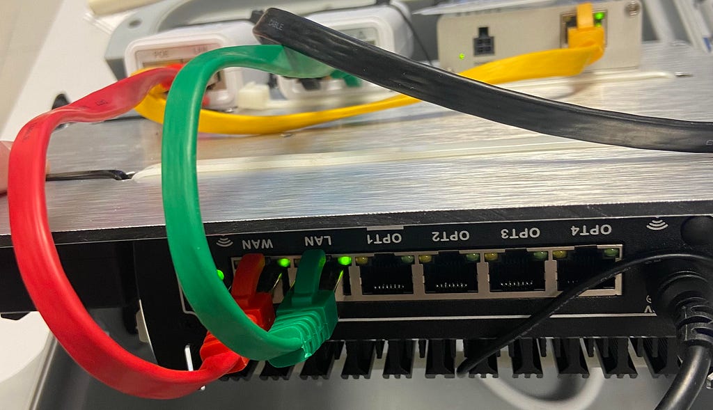 Close-up of a GEED Box network device with colored Ethernet cables connected: red and green cables in the foreground, and a yellow one in the background. Green LED indicators show network activity. The device is mounted in a technical setup, likely part of the museum’s infrastructure for GEED digital services.