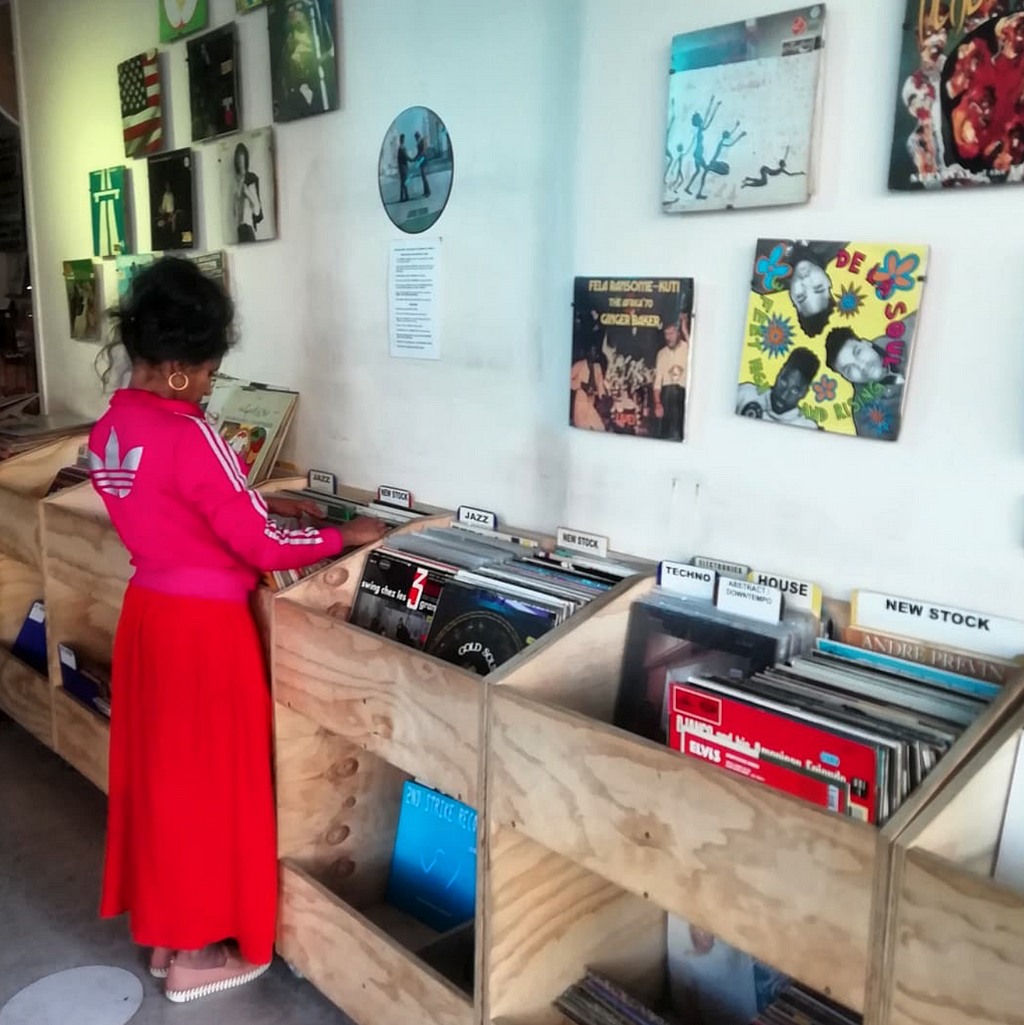 A record shop. A dark-skinned woman with black straight hair tied up, wearing a shocking pink adidas sweater and a red skirt, browses through some records