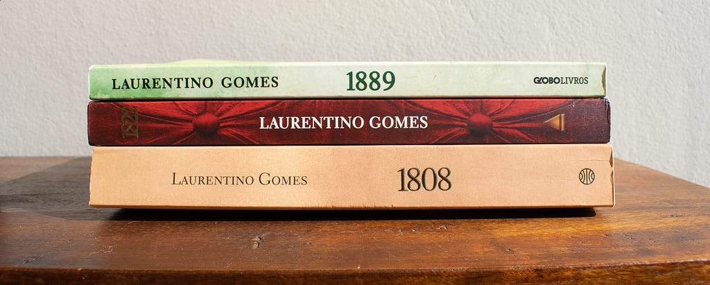 A lateral dos livros 1808, 1822 e 1889 aparece empilhad em uma estante de madeira.
