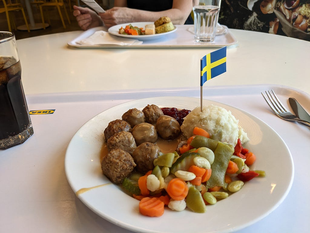 A plate of IKEA food, featuring boiled vegetables, meatballs, cream sauce, lingonberry jam, and a small Swedish flag attached to a toothpick, stabbed into the mound of mashed potatoes.