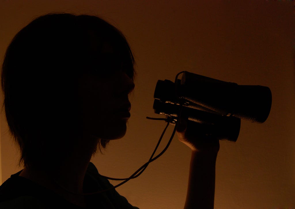 A person with shoulder length hair lit from behind, appearing like a shadow, holding a pair of binoculars
