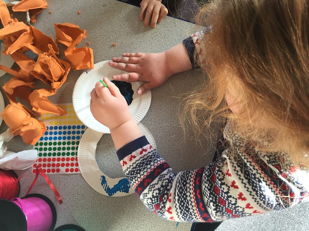 A small child painting a paper plate on top of a craft table filled with stickers, paper, ribbon.