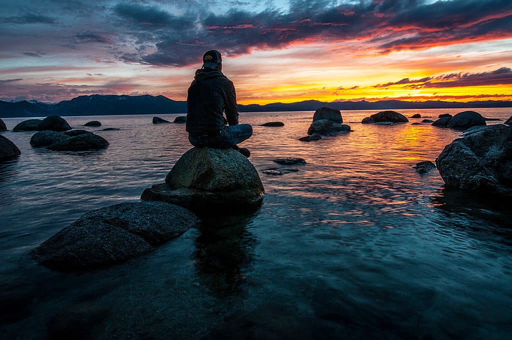 This picture shows a person sitting on a rock on a body of water. There are many stones in the water. The stones are dark in colour. The man is looking at the sky and watching the sunset. The picture of the evening and the water is mesmerizing.