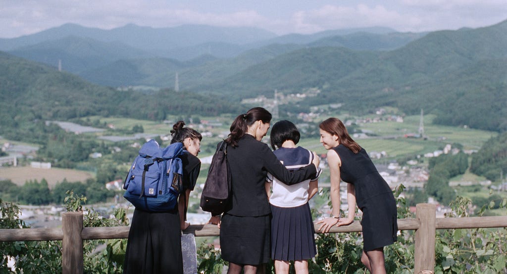 4 young women who are sisters in an embrace looking upon a vast spread of mountains and grasslands.