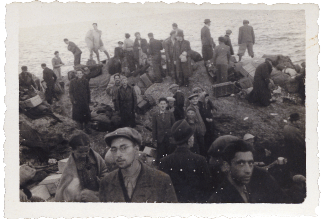 A black-and-white image of a large group of men and women standing on the rocky shoreline of an island, with suitcases and luggage around them. One young man, wearing a hat and glasses, looks to the camera.