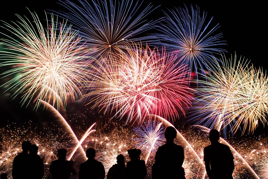 A photo of lots of huge, colorful fireworks, with the silhouettes of several people at the bottom of the image looking up at them.