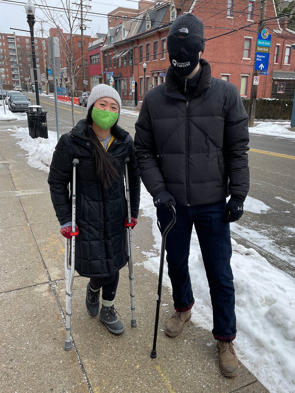 Outside on a snowy sidewalk, Thuy-Vy is posing with crutches standing next to Andrew, who is blindfolded and holding a walking cane.