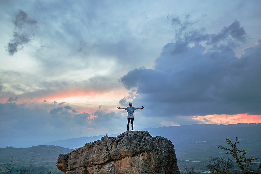 A man stands on a large rock with his hands spread eagle in front of the wide open skies and mountains