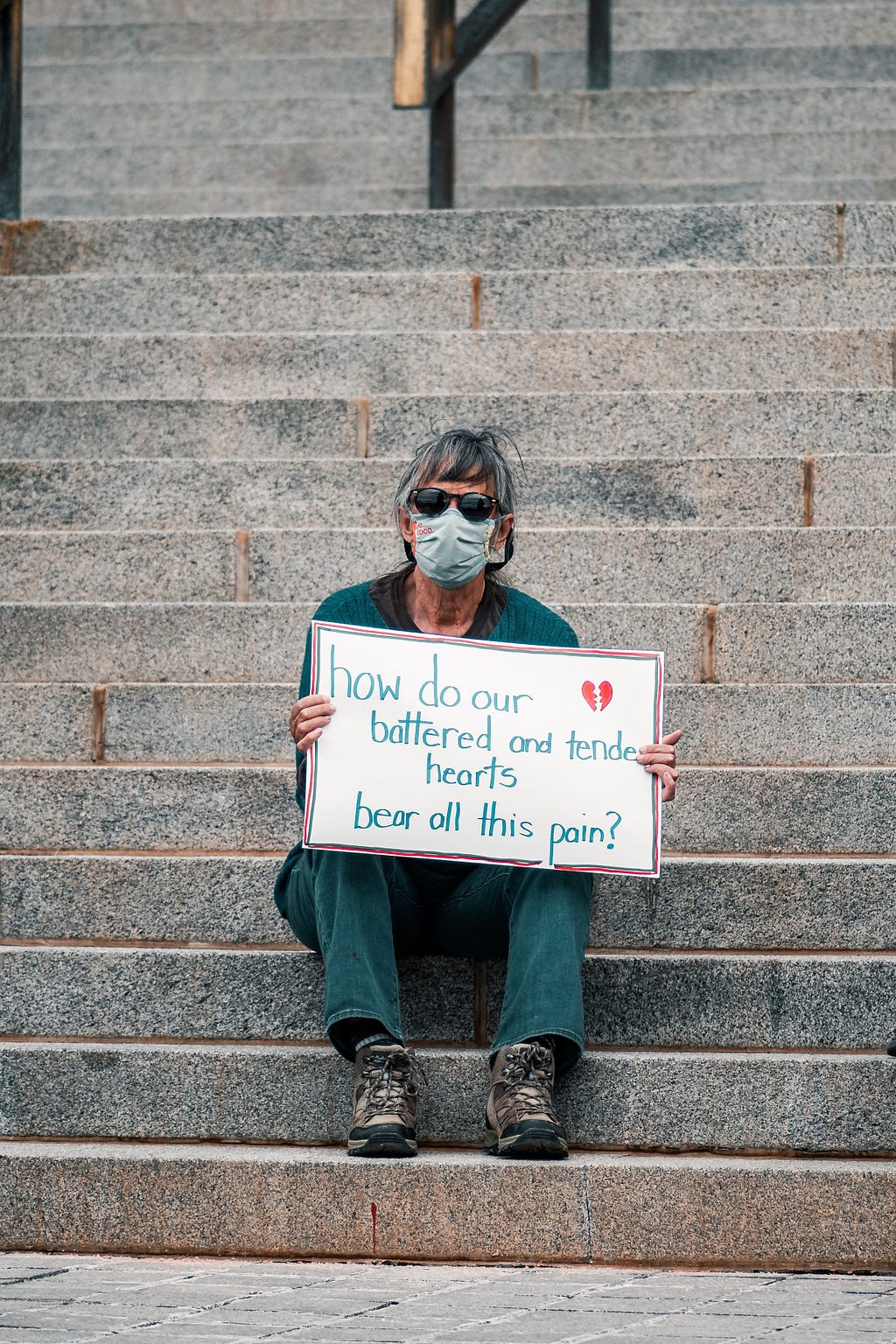 Man sitting on cement steps with a sign reading, “how do our battered and tender hearts bear all this pain?”