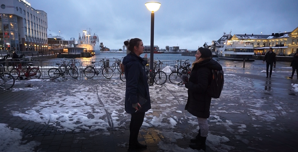Two women facing each other on a snowy street, harbour and the sea in the background
