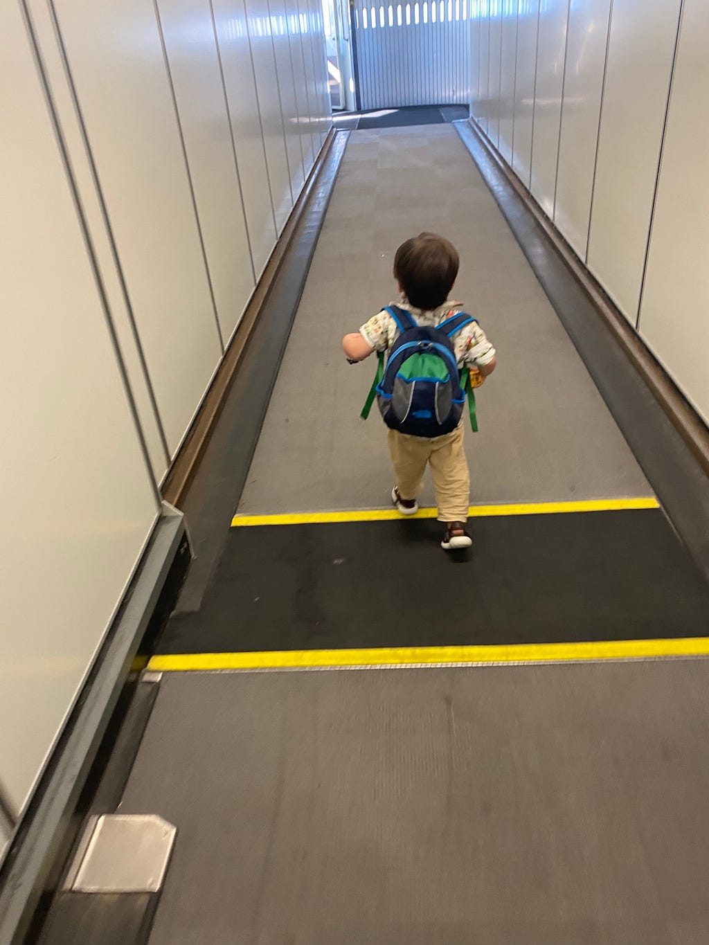 A small child seen from the back carrying a small backpack walking confidently down an airplain boarding ramp.