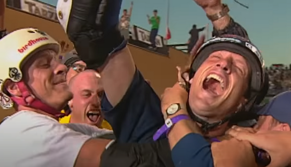 Tony Hawk celebrating, mouth open, arm in the air, surrounded by a crowd enveloping him after landing his first 900.
