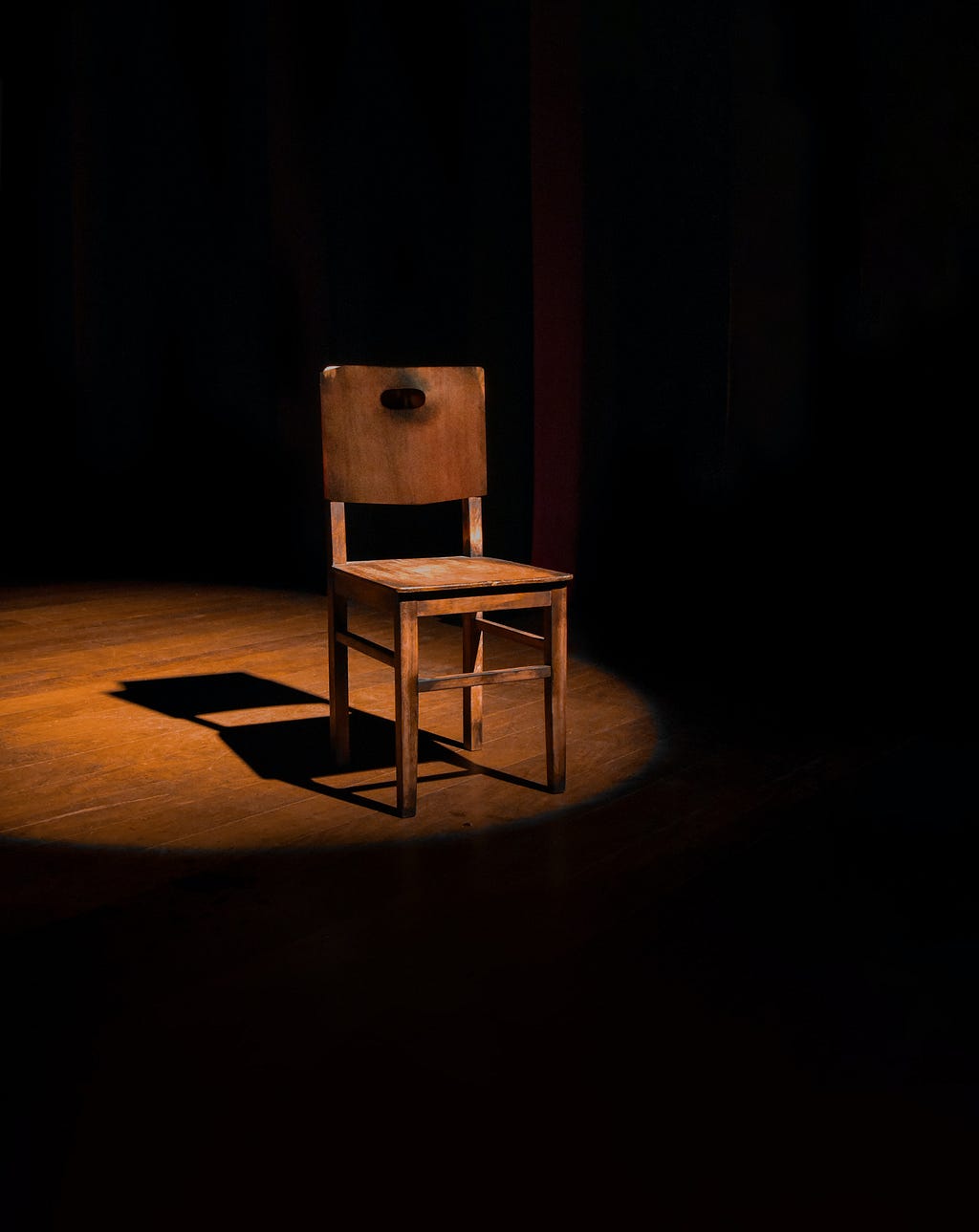 A small wooden chair in the centre of a dark room, with a spotlight cast from above.
