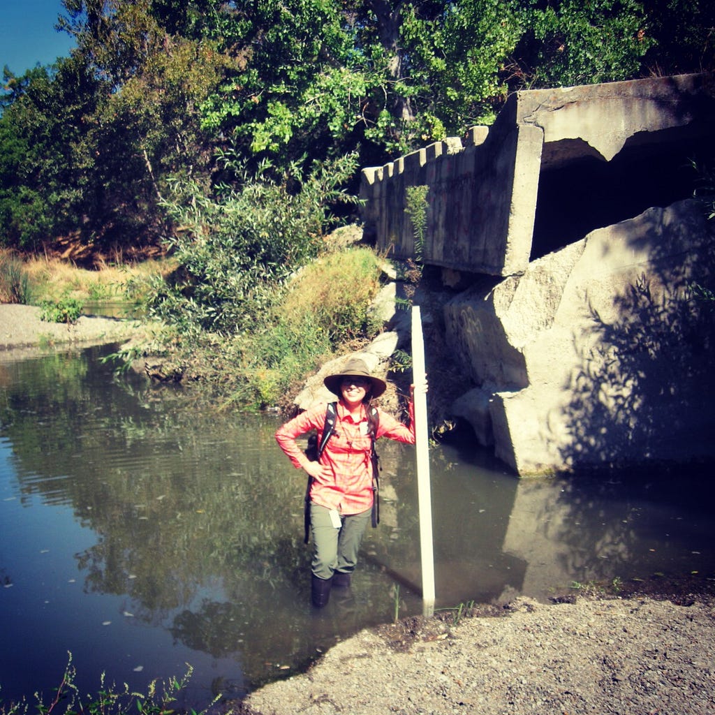 Woman wearing a hat standing in a creek in front of a concrete structure .