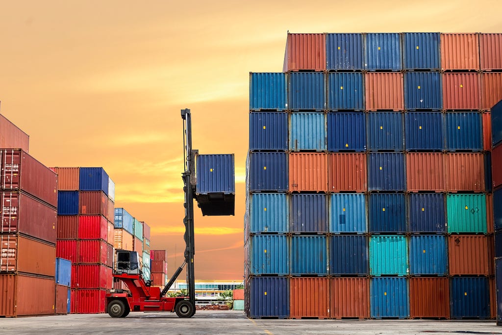 A forklift operator arranges red and blue shipping containers at a port during sunset.
