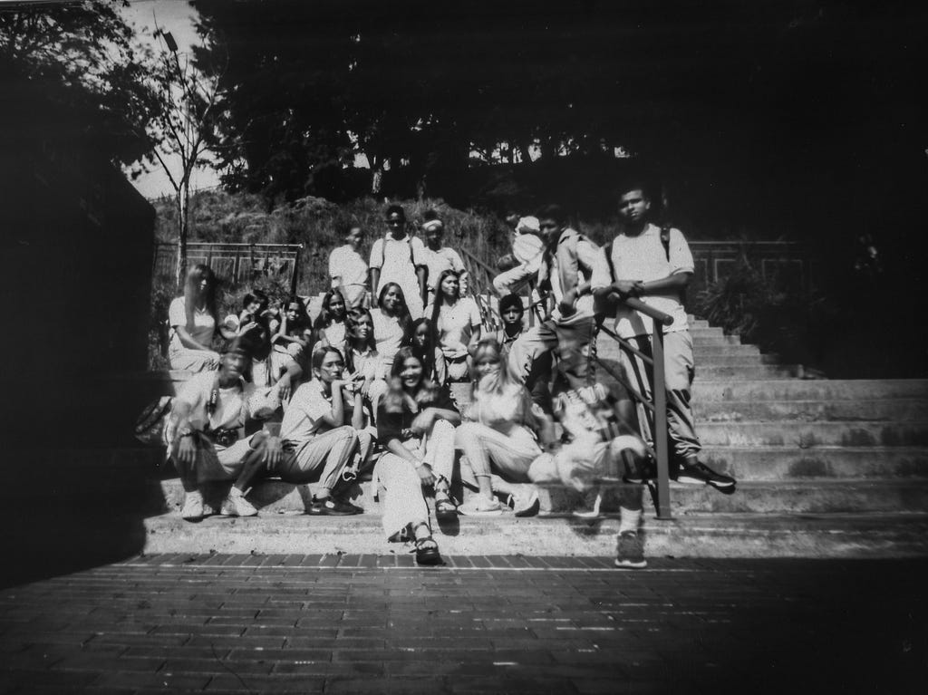 A group of around 20 teenagers sits on concrete steps outside. Image appears a bit blurry and is black and white.