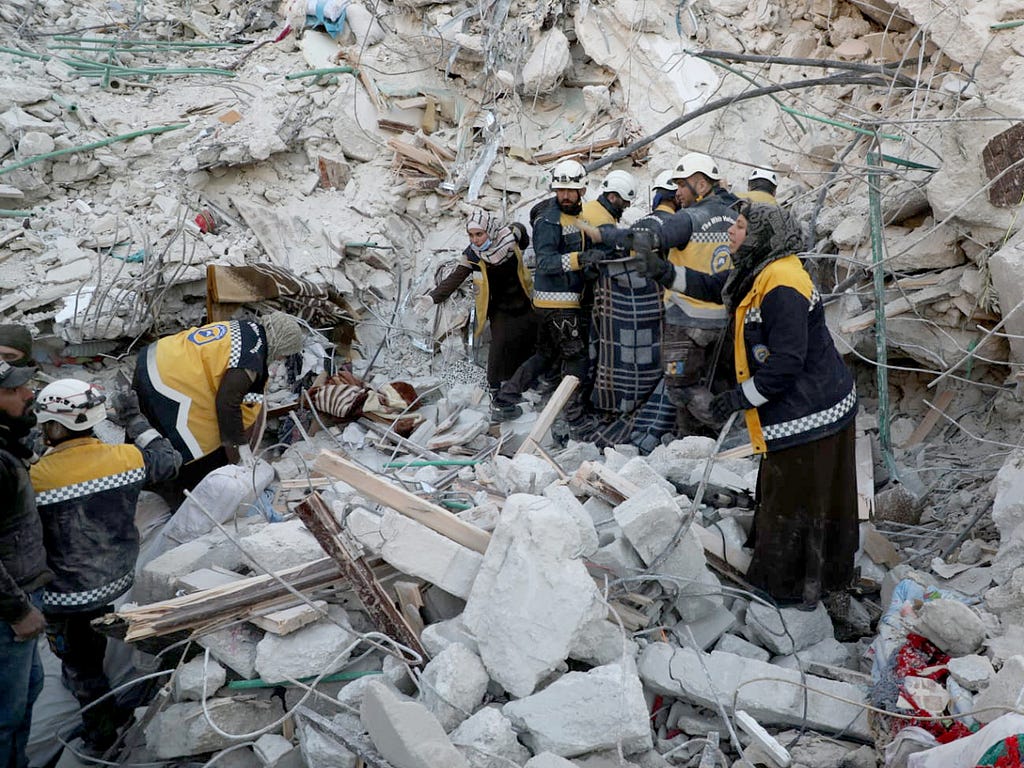 A group of people wearing yellow vests showing they are part of the White Helmets work on top and in front of rubble. The men wear white helmets. Several women wear head coverings.