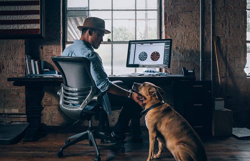 Man with hat sitting at a desk by a window, reaching out to a dog.